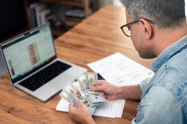 Orta yaşlı bir adam ahşap bir masada hesap tablosunu gösteren bir dizüstü bilgisayarla dolar sayıyor. Finansal planlama ve bütçe kavramı. Kişisel finans ve ev giderleri konuları için ideal 