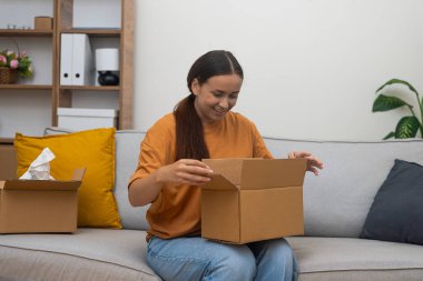 İnternetten sipariş edilen karton kutuları açan gülümseyen kadın, modern apartmanda online alışveriş tecrübesiyle tatmin olmuş bir şekilde kanepede oturuyor.