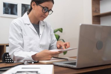 Tıbbi Çizelge: Beyaz önlüklü kadın bir doktor, modern bir klinikteki bir masada çalışırken hastanın tıbbi belgelerini inceliyor. 