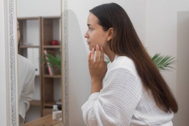 Evde güzellik ayinlerini kucaklayan bornozlu bir kadın banyo aynasında kendine bakıyor, cilt bakımını düşünüyor.. 