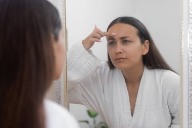 Üzgün bir kadın, banyo aynasında yansımasını inceliyor, cilt sorunları ve kırışıklıklardan rahatsız.. 