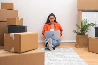 Yerleştirme işlemi devam ediyor: genç bir kadın taşınan yeni bir daireye dizüstü bilgisayarla yerleşiyor, paketlenmemiş kutuların arasına.