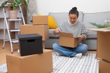 Kahverengi saçlı, neşeli yüzlü kadın kanepeye oturup modern mobilyalarla birlikte yeni bir daireye taşınmak için karton kutular topluyor.