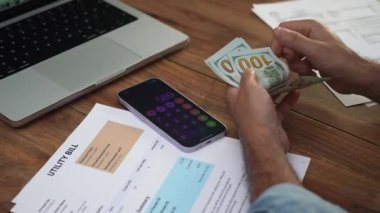 Laptop ve elektrik faturalarıyla çevrili yaşlı adam dikkatli bir şekilde masa başında dolarları sayıyor, erkek girişimci ev giderlerini karşılamak için nakit hazırlıyor.