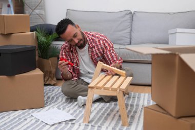 Kendi Kendini Yapmış Ev: Genç adam yeni evinde, taşınan karton kutularla çevrili, taşınma yolculuğunu kucaklayan bir DIY raf inşa ediyor.