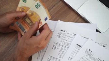Serbest çalışan adam, W-4 1040 ve W-9 vergi beyannameleri ofis belgeleriyle dolu masada oturmuş özenle parayı sayıyor.