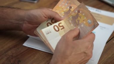 Erkek ofis çalışanı 50 avroluk banknotları titizlikle elden ele aktarıyor, adam hesaplara atıfta bulunan belgelerle ahşap masaya dokunuyor.