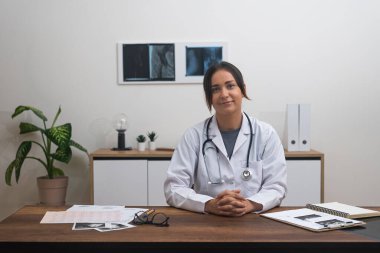 Sanal Bakım Profesyoneli: Hasta kayıtlarıyla masasında duran bir kadın doktor, çevrimiçi bir tıbbi danışma sırasında kamerayla göz teması kuruyor. 