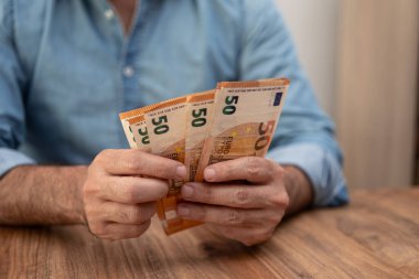 Avro banknotlarını elinde tutan erkek girişimci, ahşap masada oturan pahalı ofis kiralama hizmetleri için ev sahibine ödeme yapmak üzere dikkatlice para sayıyor.