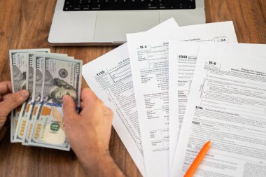Serbest çalışan adam, işyerinde dizüstü bilgisayarla oturan çeşitli vergi formlarını doldurmak için gelir hakkında ayrıntılı finansal bilgiler hazırlayan dolar banknotlarını sayar.