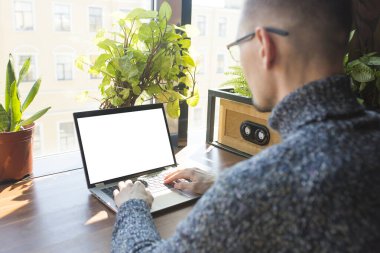 Büyük pencerenin yanındaki ahşap masada boş beyaz masaüstü ekranlı dizüstü bilgisayar kullanan gözlüklü iş adamının görüntüsü..