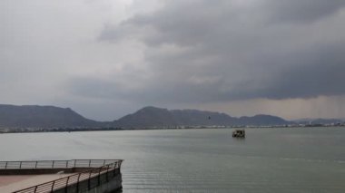 Yağmurlu bir günde gökyüzünde gri bulutlar ve arka planda tepeler olan bir su kütlesinin görüntüsü. Ajmer 'de Ana Sagar Gölü' nün manzara görüntüsü.