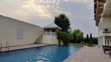Sawai Madhopur, Hindistan 8 Haziran 2024: Gündüz vakti çekilmiş bir oteldeki yüzme havuzu, ağaçlar ve sandalyelerin hızlandırılmış görüntüleri. Mavi, havuzu yansıtan şehir mimarisi manzarası ortamı