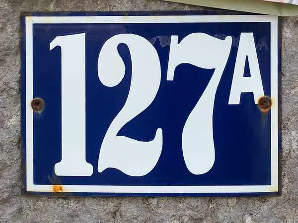 Yüz yirmi yedi ve harf A ya da 127 A, mavi metal plakalı, beyaz ev numarası ile, kapat. Arapça rakamlar: bir, iki, yedi. Taş duvar arka planında, sokakta geleneksel rakamlar.