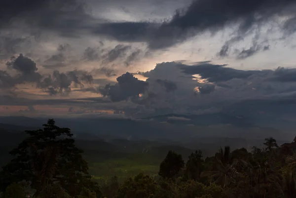 Indonesian landscape with rainy sky on sunset - dark blue clouds with orange, yellow sunbeams over hazy mountains, smoky green valley with rice plantations, tropical forest on Bali, Munduk village.