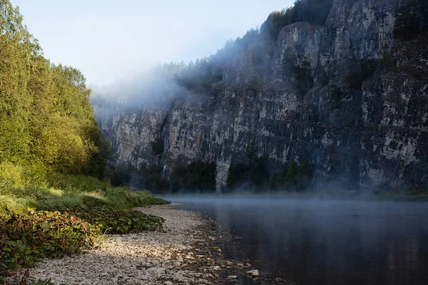 Majestic morning summer landscape in river canyon with rocky cliff in blue shadow and fog on river with white haze flow above water, lush green forest on shore in bright golden sunlight.
