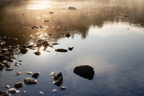 Golden sunbeams and ripple glare on calm river water surface with fluffy yellow fog, blue reflection of sky, stones on shore in early morning. Bright, warm, quiet sunrise background, detail, texture.