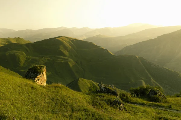 stock image Majestic summer mountain landscape - bright green mountains ridges, slopes in golden light of early morning, soft mist, lush green meadow, boulder on ledge, panorama view. Amazing trip in Dagestan.