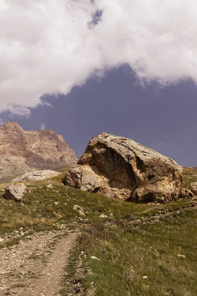 Summer mountain landscape - big yellow boulder with cracks on country road, green grass meadow, high pink rocky cliff away in sunny day with blue sky, white clouds. Travel in Dagestan, vertical.