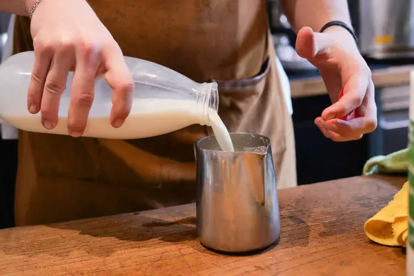 A person is adding milk to a measuring cup as part of a recipe. The measuring cup is made of wood and placed on a hardwood table. The persons fingers and thumbs are visible in the gesture