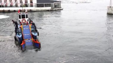 Mürettebatlı küçük bir tekne limanda yüzen çöpleri topluyor ve su temizleme çalışmalarına katkıda bulunuyor.