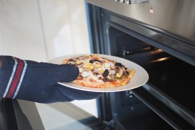 Mutfak sahnesinde bir kişi fırından taze bir pizzayı dikkatlice çıkarmak için fırın eldiveni kullanır.