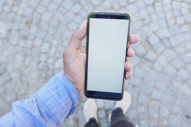 Mavi tişörtlü bir kişi dışarıda kaldırım taşı arka planda dikilirken boş ekran akıllı telefon tutar.