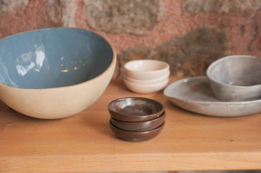 Kırsal seramik kaseler ve tabaklar ahşap bir rafta dizilmiş, bir mutfakta veya iç mekanda taş bir duvara karşı dizilmiş.