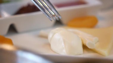 Bir çatalın yakın çekim çerçeveleri peyniri kaldırıyor ve kremalı bir tatlı ve arka planda başka yiyecekler var..