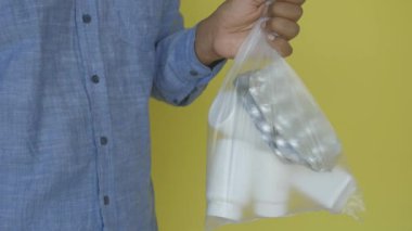 Bir adam, parlak sarı arka planda çeşitli ilaçlar içeren bir çanta tutarken görülüyor.