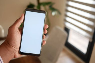 Görüntü, beyaz ekranlı akıllı bir telefonu tutan çağdaş bir iç mekanda bir eli gösteriyor.