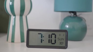 Sabah 7: 10 'u gösteren dijital alarm saati, vazo ve lamba yanında modern beyaz bir masaya kurulmuş..