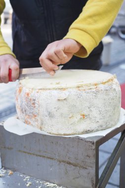 Uzman eller, açık hava pazarında keskin bir bıçakla büyük bir peynir çarkını kesiyor. Detaylı bir yakın çekim sırasında yakalanmış.