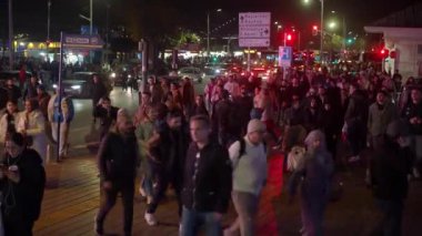 Şehir kavşağında yoğun yaya trafiği ve hareket halindeki araçlarla kalabalık bir gece caddesi..