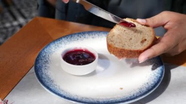 Bir insan kahvaltı masasında otururken bıçak kullanarak taze ekmeğe reçel sürer..