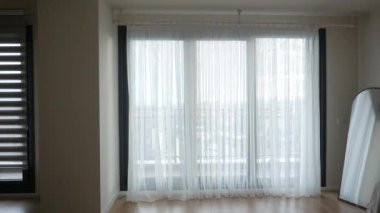 Büyük pencereli parlak oda, ahşap zemin, yeşil bitki ve yuvarlak ayna..