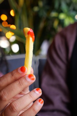 Yakın plan, zengin, keskin ketçapa batırılmış patates kızartmasını kavrayan bir eli gösteriyor.