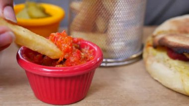 Patates kızartmasını canlı domates sosuna batıran bir el. Arka planda bir yemek görünüyor.