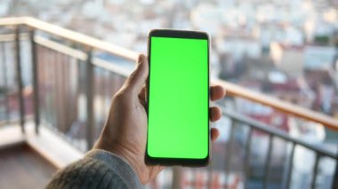 Bir kişi canlı yeşil ekranlı akıllı bir telefon tutar, manzaralı bir arkaplan üzerine kuruludur.