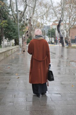 Zarif kahverengi ceketli şık bir figür ve sakin bir parkta rahatça geziniyor.