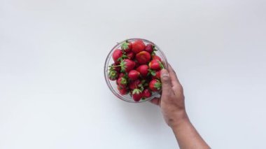 Bir el, temiz, beyaz bir yüzey üzerinde olgun çileklerle dolu bir kase tutar.