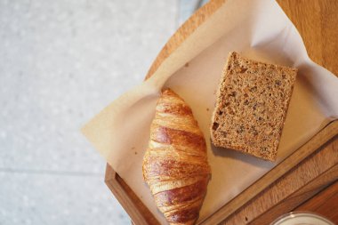 Sıcak ve pullu bir kruvasan, ahşap tepside duran tahıllı ekmeğin yanında.