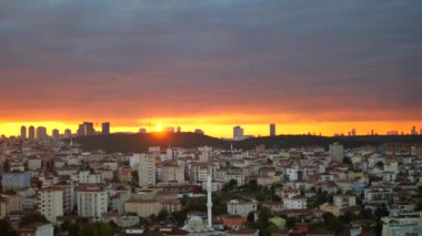 Çarpıcı ve dramatik bir günbatımı, geniş bir şehrin ufuk çizgisi üzerinde canlı turuncu renkler bırakır.