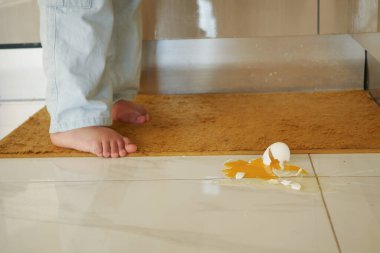 Serin mutfak zemininde çıplak ayakla duran küçük bir çocuk, kırık bir yumurtanın yanında merakla bakıyor.