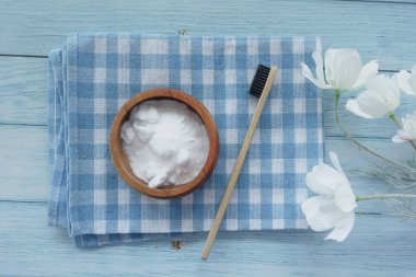 Mavi kareli kumaş üzerinde ahşap bir kase kabartma tozu, bambu bir diş fırçası ve cam bir kavanoz sergilenir.