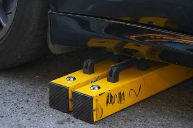 Parlak sarı bir tekerlek kilidi belirgin bir şekilde sergileniyor, yakınlarda park edilmiş parlak siyah bir arabanın altında korunuyor.