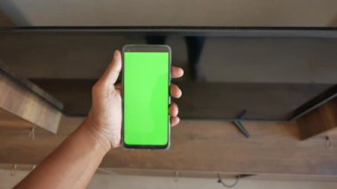 Televizyonun yanında yeşil ekranlı akıllı bir telefon tutan bir kişi var.