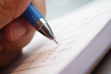 Yakından bakınca mavi bir kalemi tutan ve bir kağıt parçasına dikkatlice yazan bir el görülüyor.