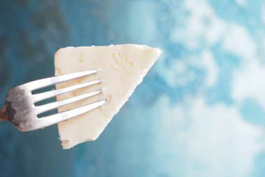 Bir çatal tarafından kaldırılan, canlı bir mavi arkaplan üzerine kurulmuş, baştan çıkarıcı bir peynir parçası..