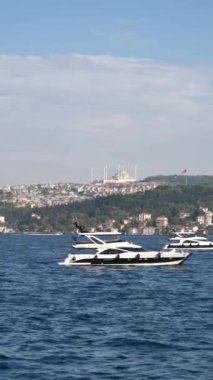 Gemiler sakin sularda süzülür, çarpıcı bir şehir silueti uzaklardan görülebilir.